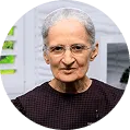An older woman with short gray hair and glasses, wearing a dark patterned top, is looking at the camera. The bright background features white elements and green blurred foliage, making her an ideal fit for a welcoming Become A Patient banner.