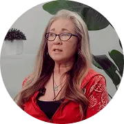 A woman with long blonde hair and glasses, wearing a red patterned shirt, sits indoors in front of a white wall with green plants beside a Patient Banner.