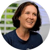A woman with short brown hair is smiling and wearing a blue shirt. She is indoors with blurred white blinds and greenery in the background, perfect for a healthcare patient banner.